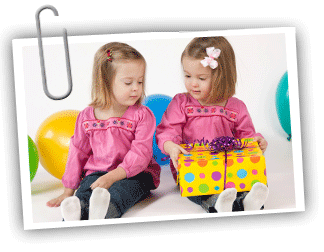 Two twin girls sitting on the floor surrounded by balloons with the twin on the right holding a present